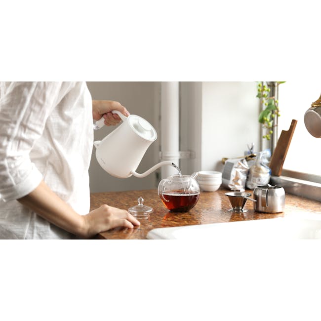 BALMUDA The Pot - White - 2 - White teapot, brown countertop, person pouring tea, clear glass teacup, dark amber tea, white