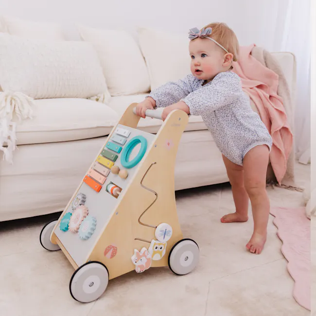 Bubble Wooden Activity Play Walker - 7 - Baby in floral pajamas, standing on carpet, holding a wooden walker, colorful toys