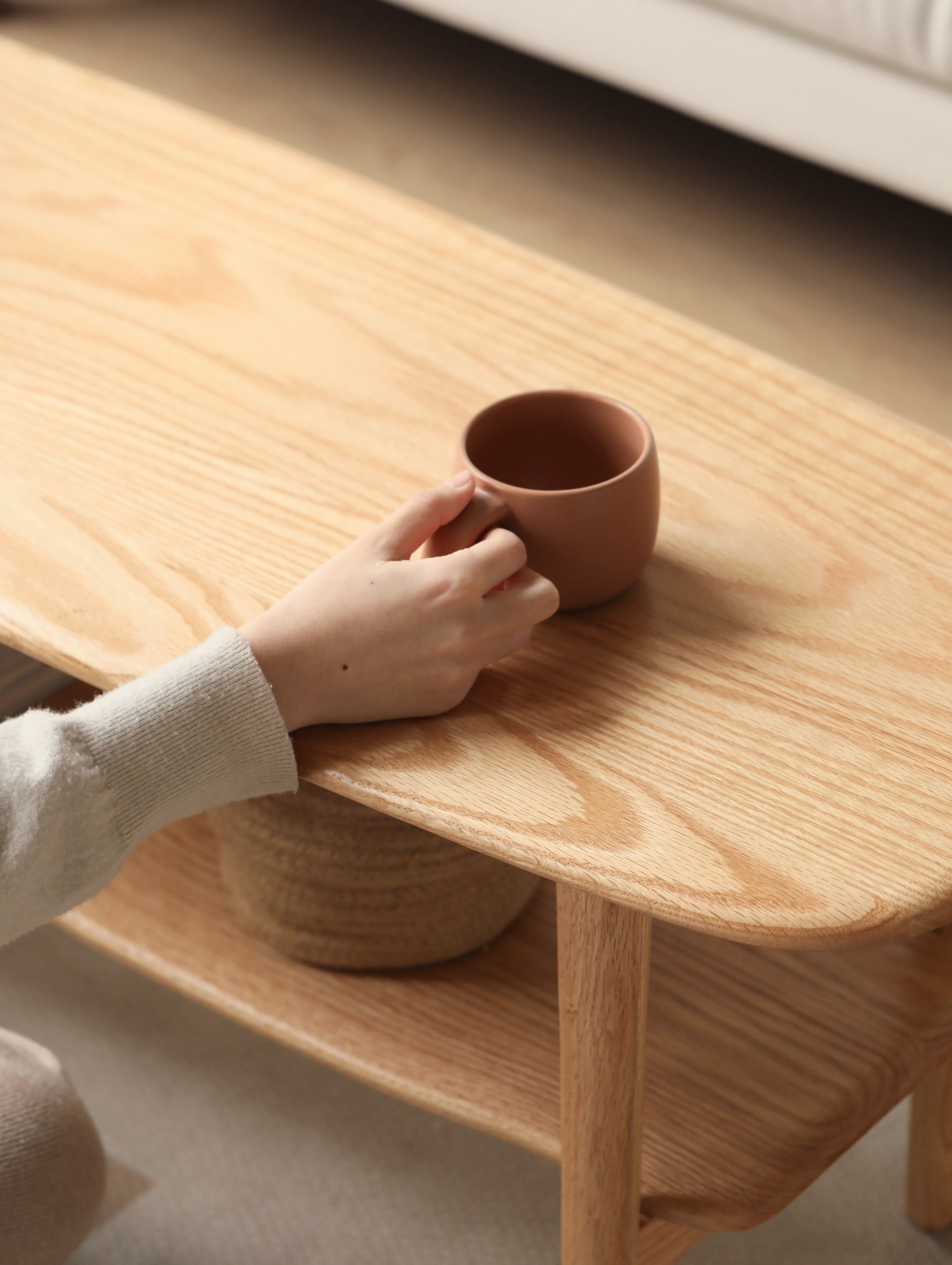 Nigel Narrow Coffee Table 1m - 9 - Hand holding brown ceramic mug, wooden table surface, beige sweater sleeve, wooden shelf