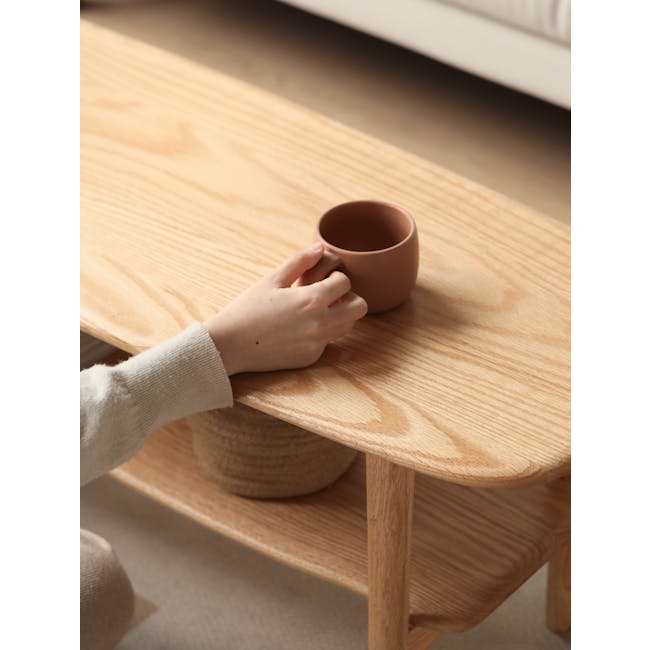 Nigel Narrow Coffee Table 1m - 9 - Hand holding brown ceramic mug, wooden table surface, beige sweater sleeve, wooden shelf