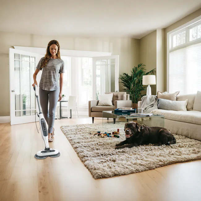 Shark Steam and Scrub Mop - 2 - Woman vacuuming living room, dog playing on rug, beige sofa, glass coffee table, white lamp
