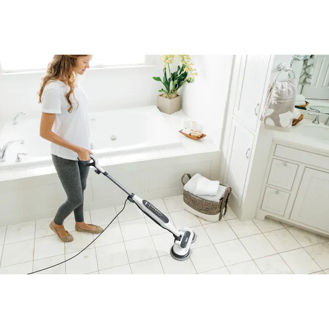 Shark Steam and Scrub Mop - 4 - White bathroom, woman vacuuming, gray leggings, beige shoes, white towel, beige basket