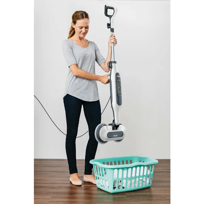 Shark Steam and Scrub Mop - 6 - Woman holding Shark vacuum cleaner, standing next to turquoise laundry basket, gray shirt