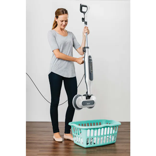 Shark Steam and Scrub Mop - 6 - Woman holding Shark vacuum cleaner, standing next to turquoise laundry basket, gray shirt