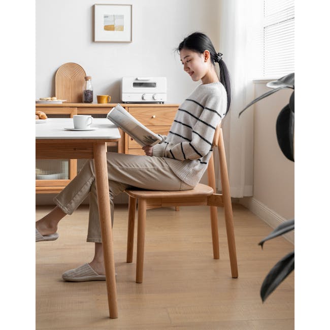 Solis Dining Chair - Oak - 3 - Sitting on wooden chair, reading magazine, light gray sweater, beige pants, light brown