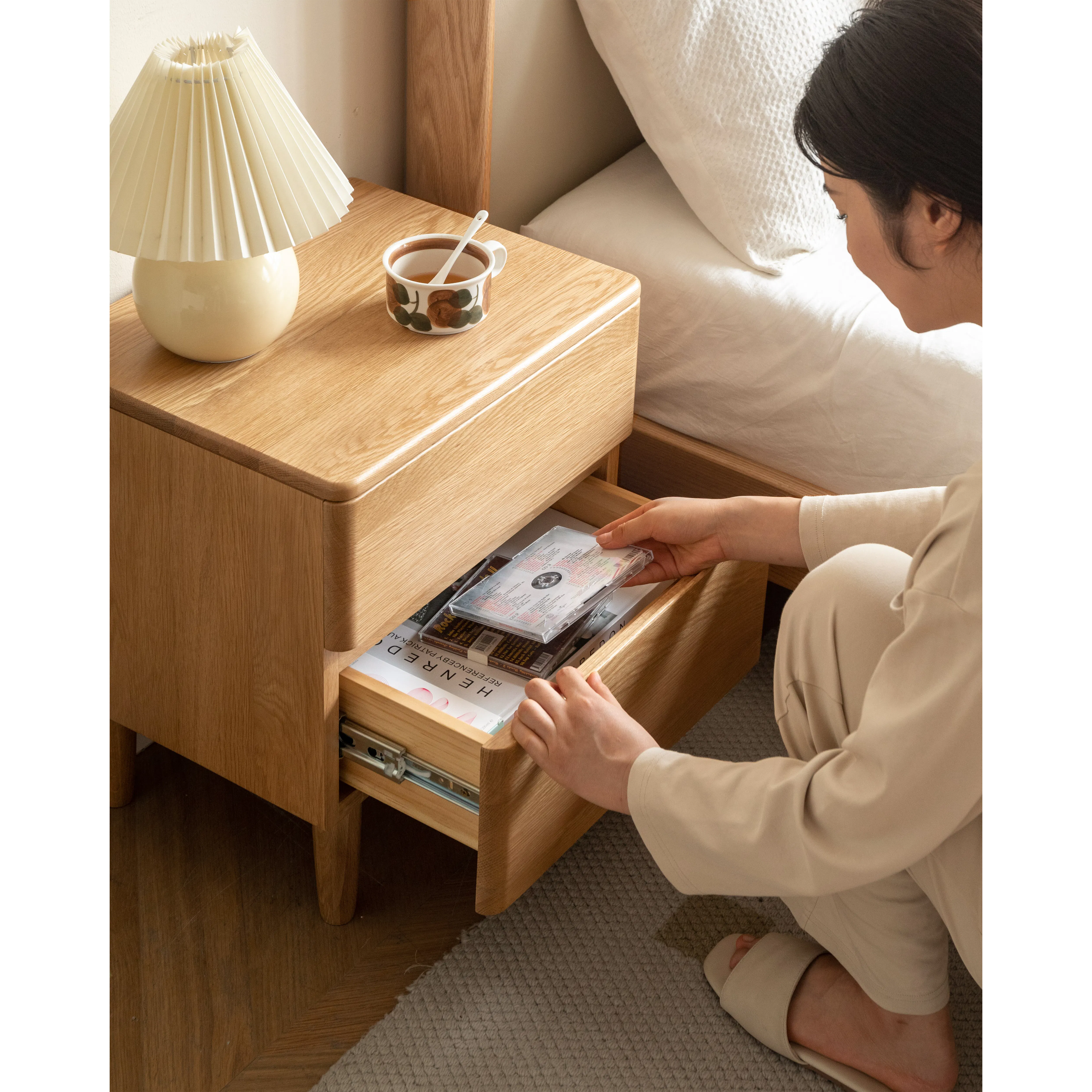 Natalia Bedside Table - 18 - Wooden nightstand, beige lamp, cup with spoon, wooden drawer, person's hand, beige pants, beige