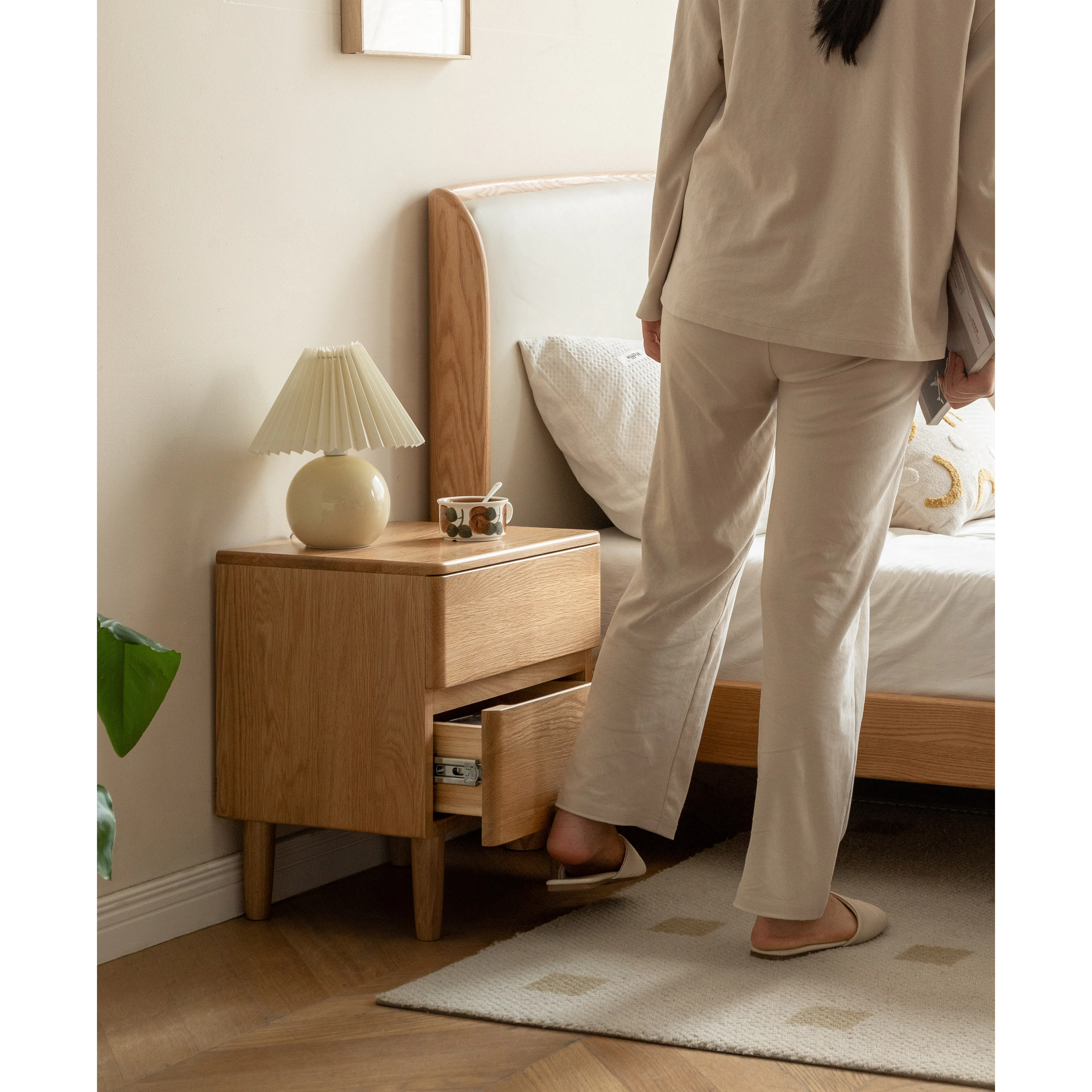 Natalia Bedside Table - 19 - Wooden nightstand, beige lamp, white bed, beige pants, beige shoes, person standing, wooden