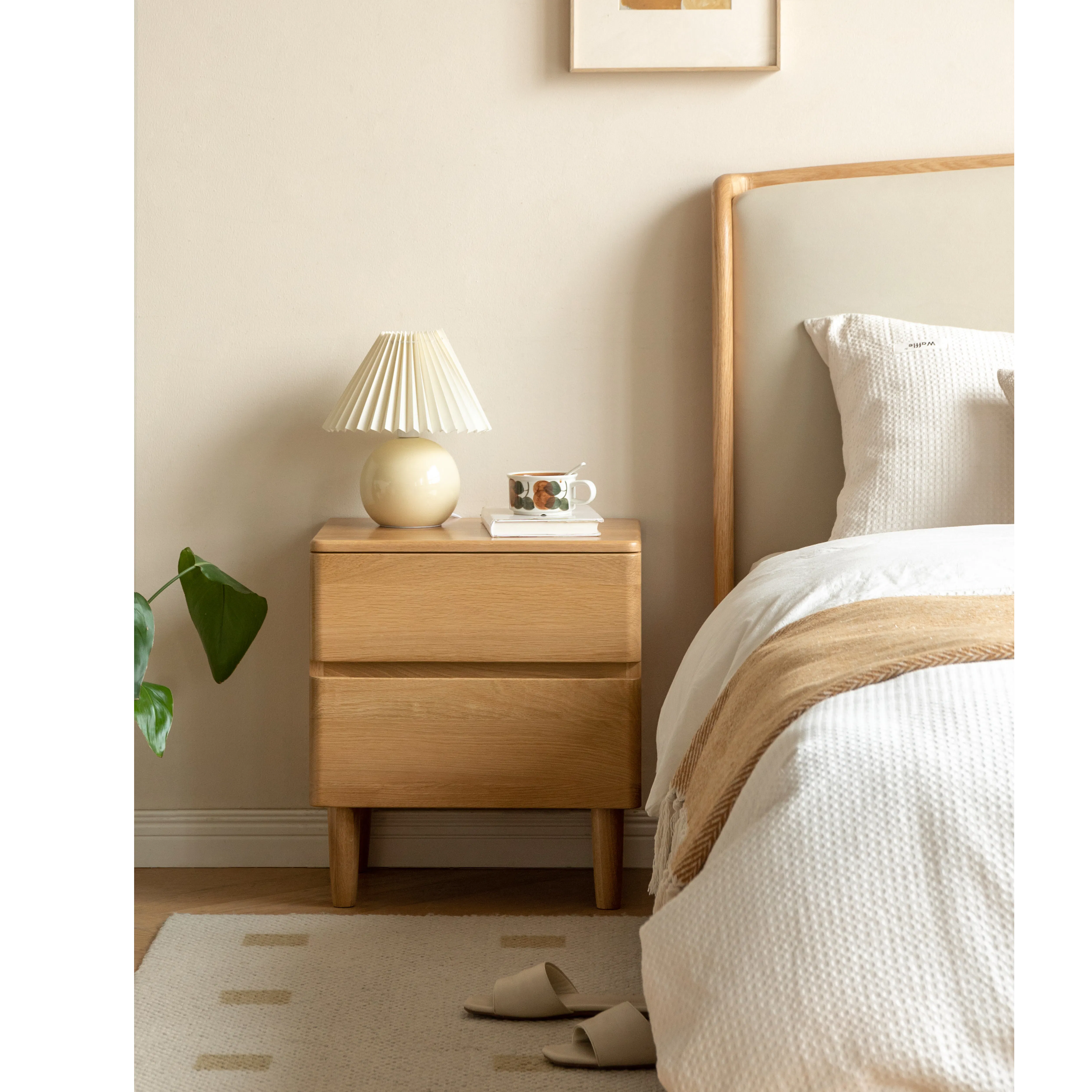 Natalia Bedside Table - 12 - Wooden nightstand, beige lamp, white mug, green plant, wooden headboard, white bedding, beige