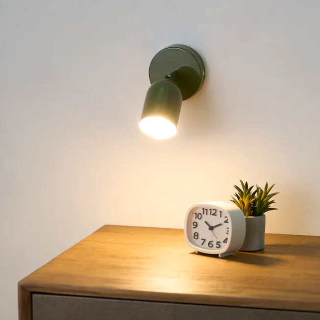 Hetta Wall Light - Green - 2 - Green wall-mounted light fixture, wooden desk, white analog clock, potted plant, warm