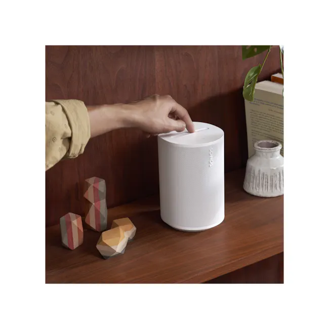 Sonos Era 100 - Black - 2 - White Sonos speaker, wooden shelf, hand interacting with device, geometric stones, natural wood