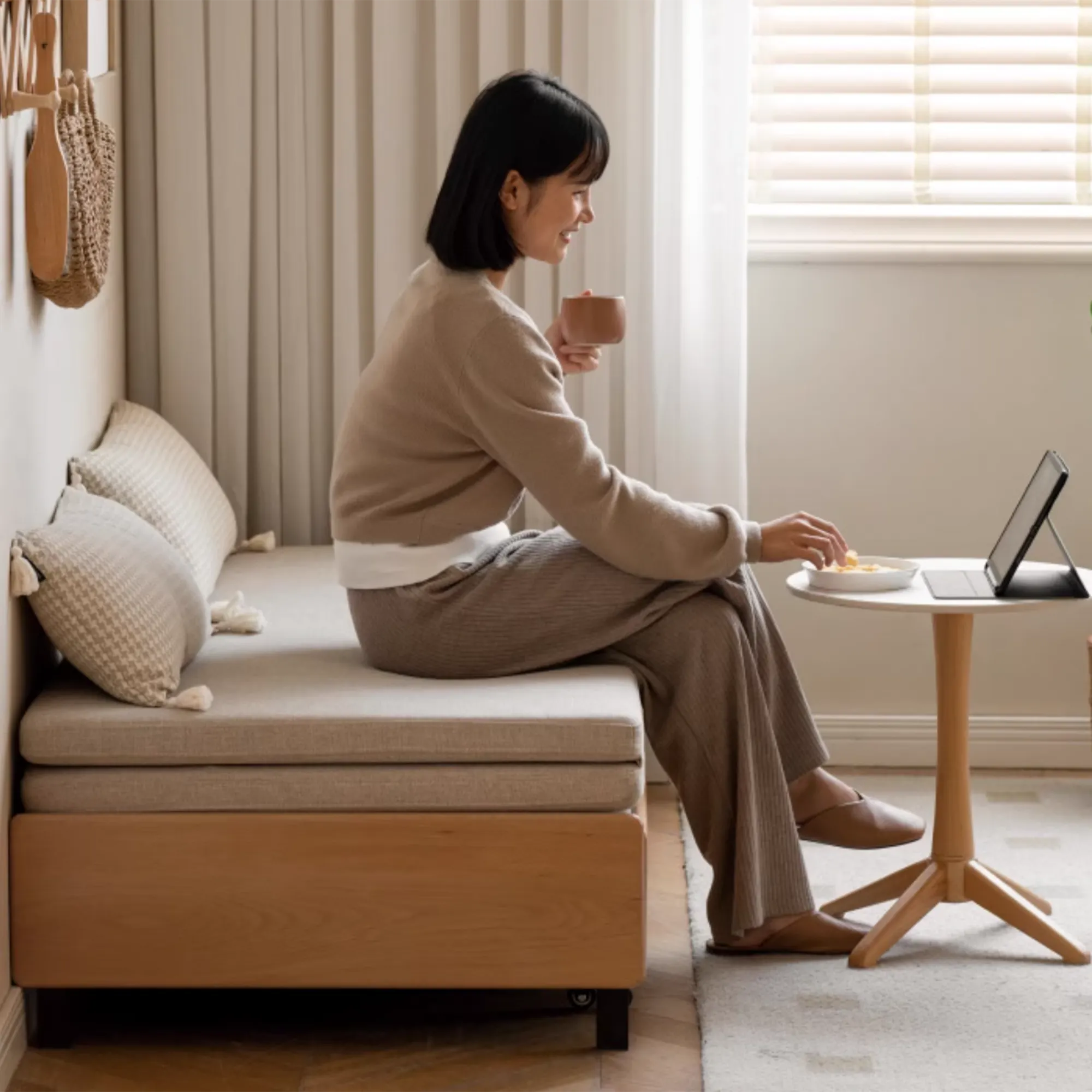 Yoke Sofa Bed / Bench 1.5m - Gravel White - 10 - Sitting on a cozy bed, holding a cup, smiling, wearing a beige sweater