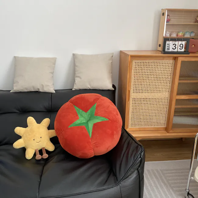 Tomato Plush Cushion - 3 - Comfortable black leather couch with two beige pillows, Large red tomato-shaped plush toy with