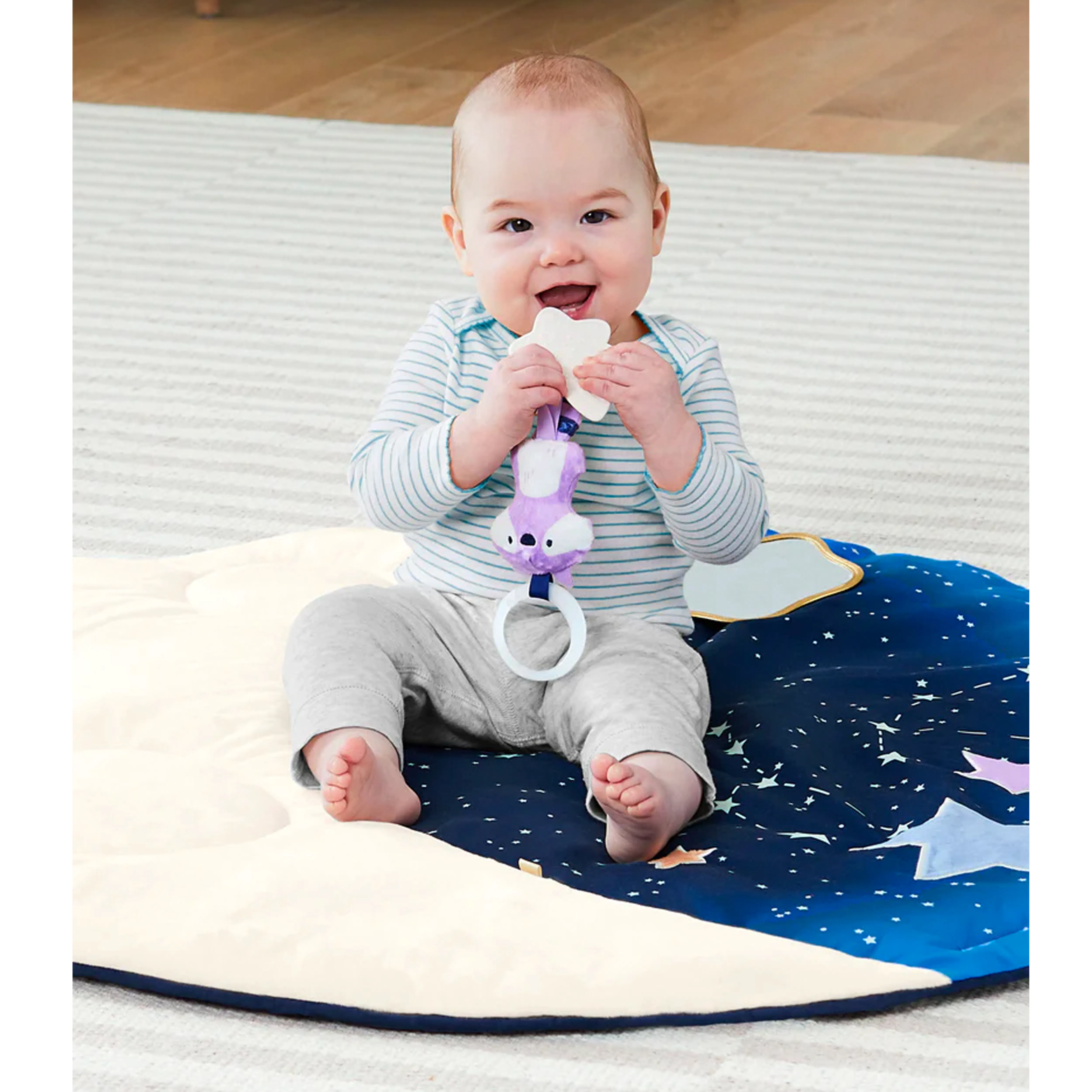 Skip Hop Activity Gym - Celestial Dreams - 13 - Baby sitting on a star-patterned blanket, holding a white toy, wearing a