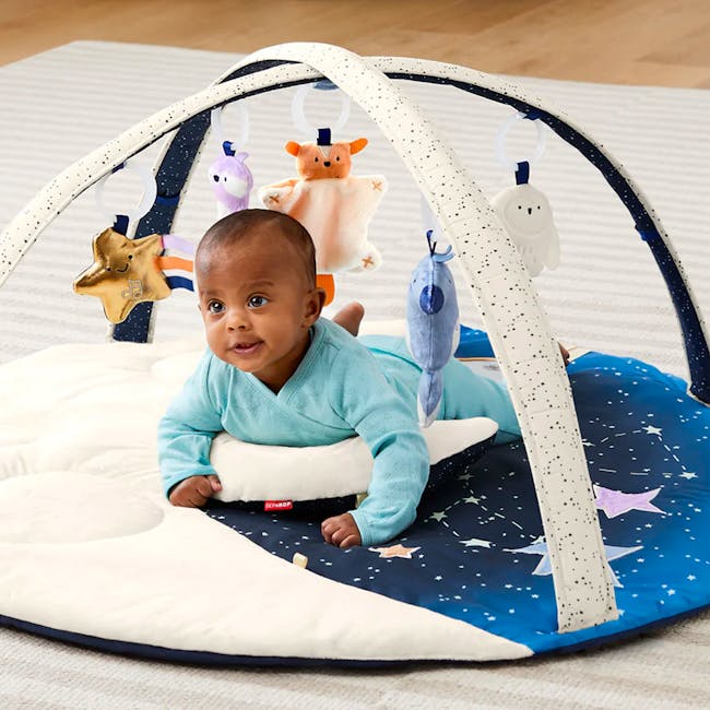 Skip Hop Activity Gym - Celestial Dreams - 11 - Baby in blue outfit lying on a starry play mat, surrounded by plush toys