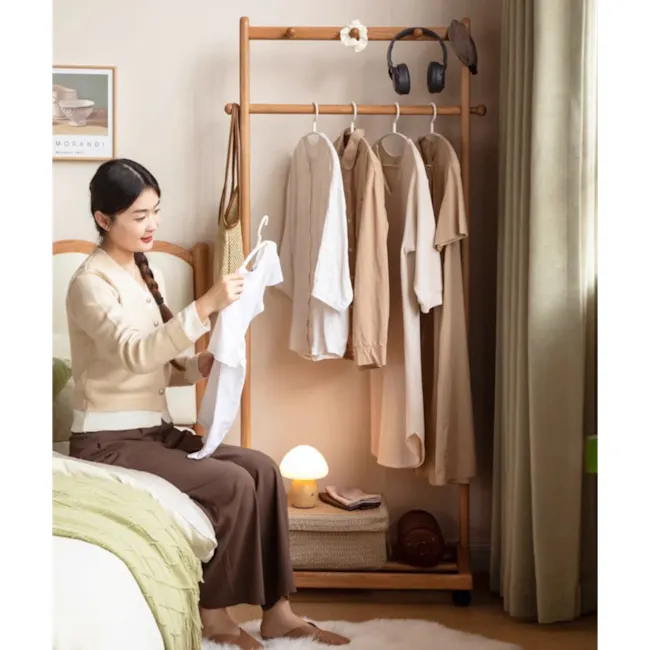 Pensa Clothes Rack - Oak - 5 - Woman sitting on bed, examining white shirt, beige clothing rack with various garments, soft