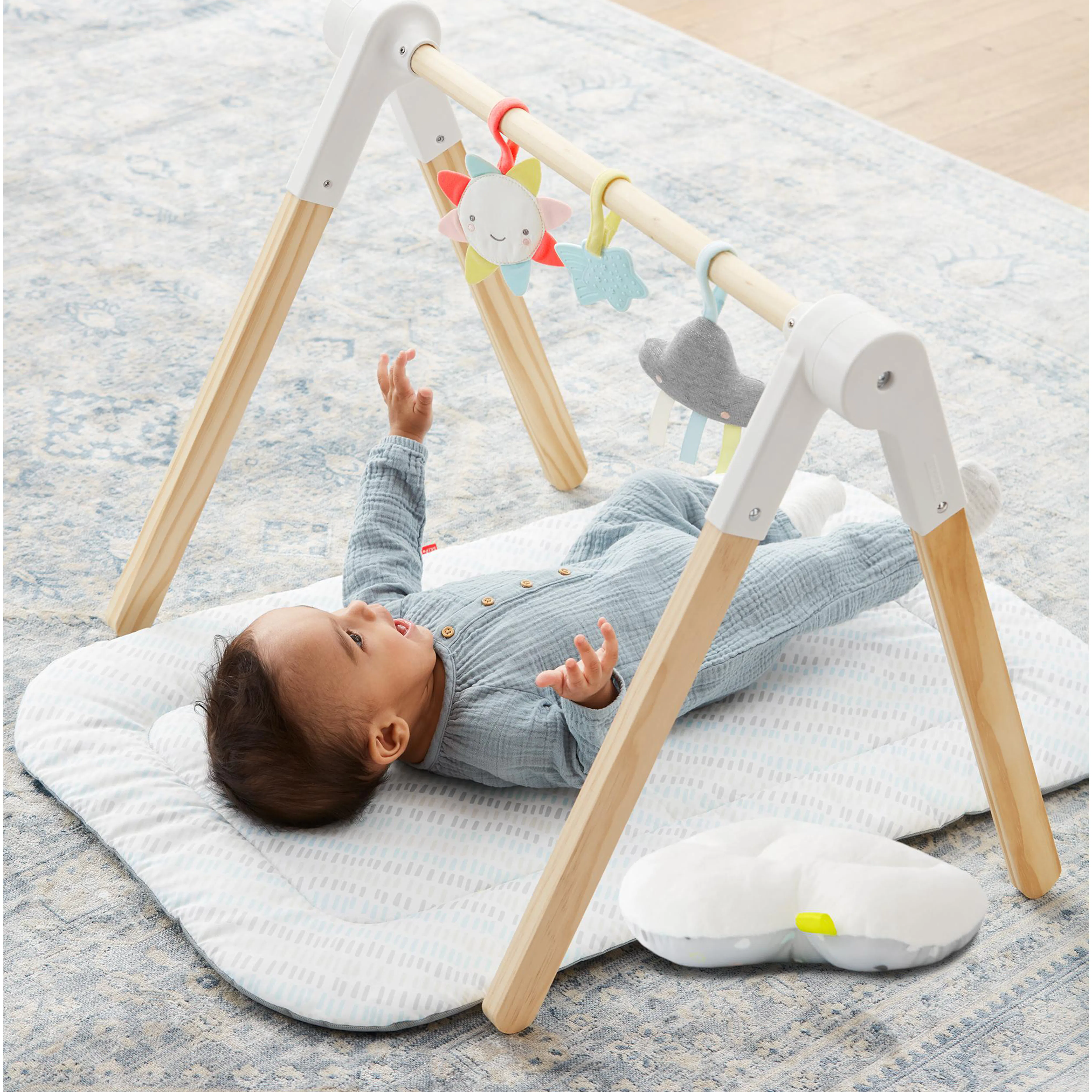 Skip Hop Wooden Activity Gym - Silver Lining Cloud - 10 - Baby lying on a soft, patterned rug, wearing a blue outfit
